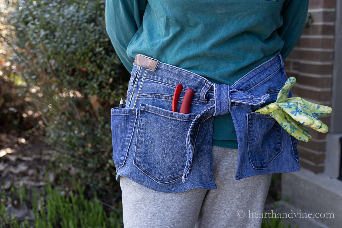 A person wearing the jean gardening apron with pruners, garden gloves and plant labels in the pockets.