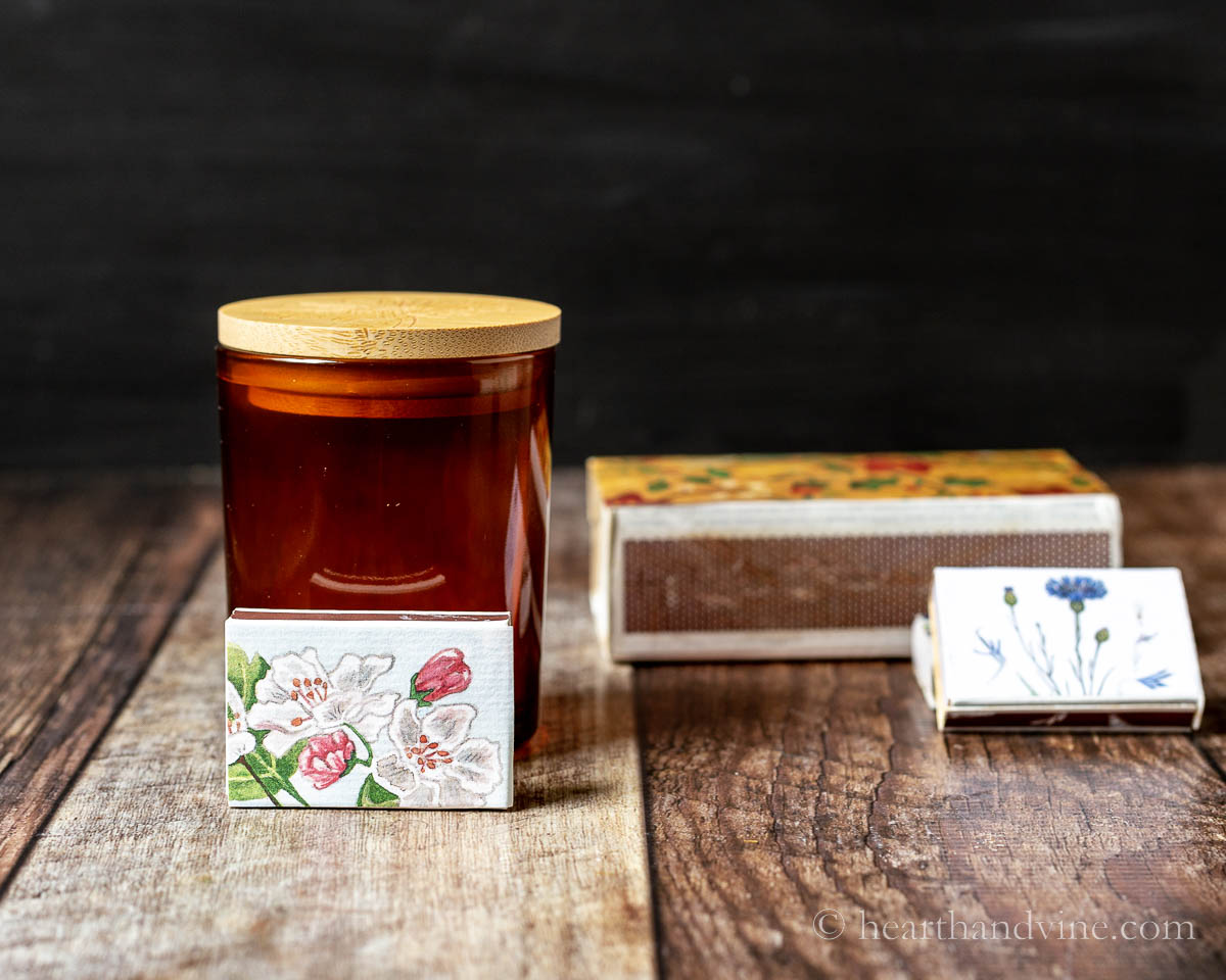 A glass candle container with a wooden lid next to a small floral matchbox and a large one and a couple of small boxes to the right.