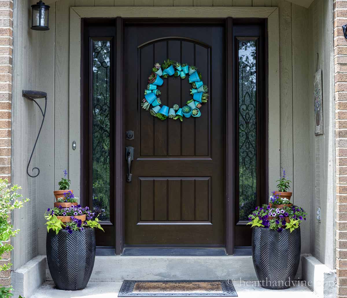 Front door view with a clay pot wreath in shades of blue and two large black pots flanking the door with stacked pots filled with annuals in shades of purple and two chartreuse sweet potato vines.