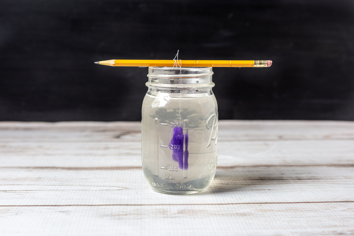 Purple pipe cleaner shape in the middle of a mason jar filled with crystal solution hanging from a pencil with thread.