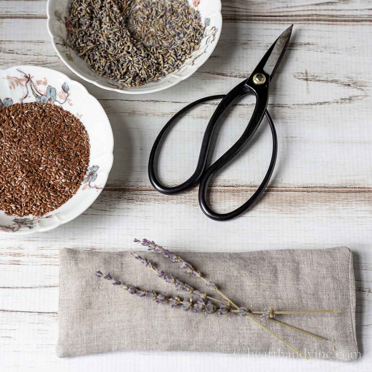 Linen eye pillow with dried lavender and bowls of flaxseed and lavender buds next to a pair of scissors.