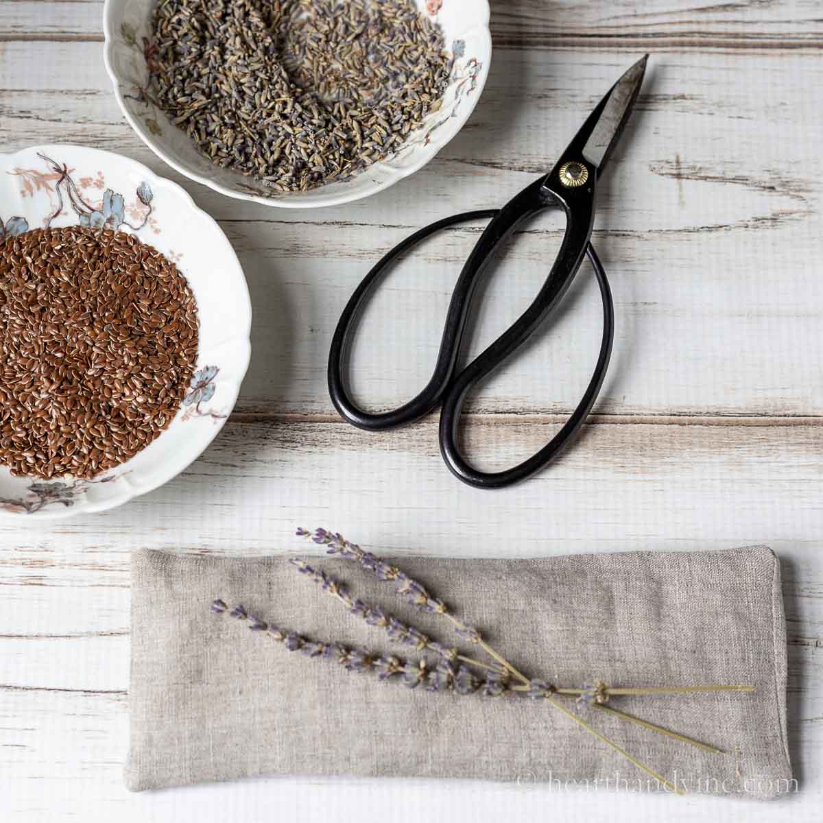 Linen eye pillow with dried lavender and bowls of flaxseed and lavender buds next to scissors.