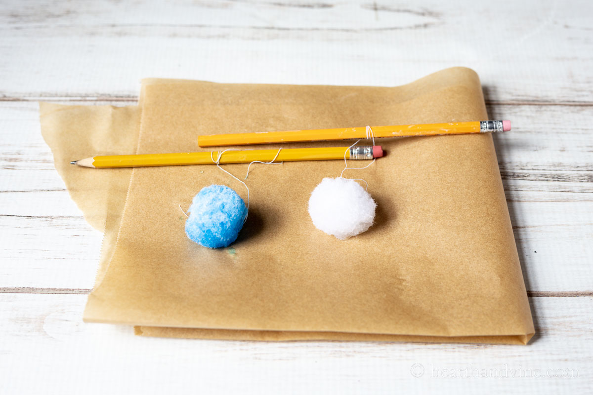 Two pom poms. One blue and one white drying on parchment paper tied to pencil with thread.
