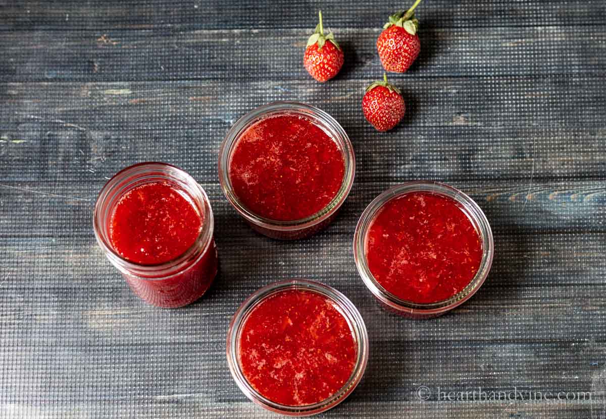 Aerial view of open jars of strawberry jam and three fresh strawberries on the top.
