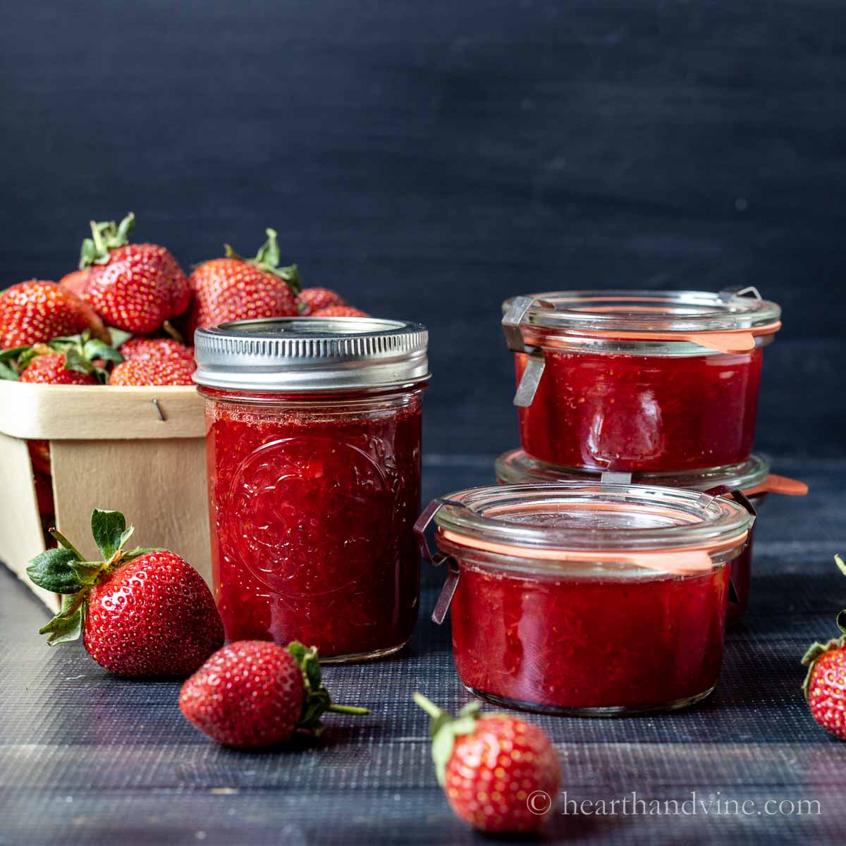 Jars of strawberry jam surrounded by fresh strawberries and a pint of berries.