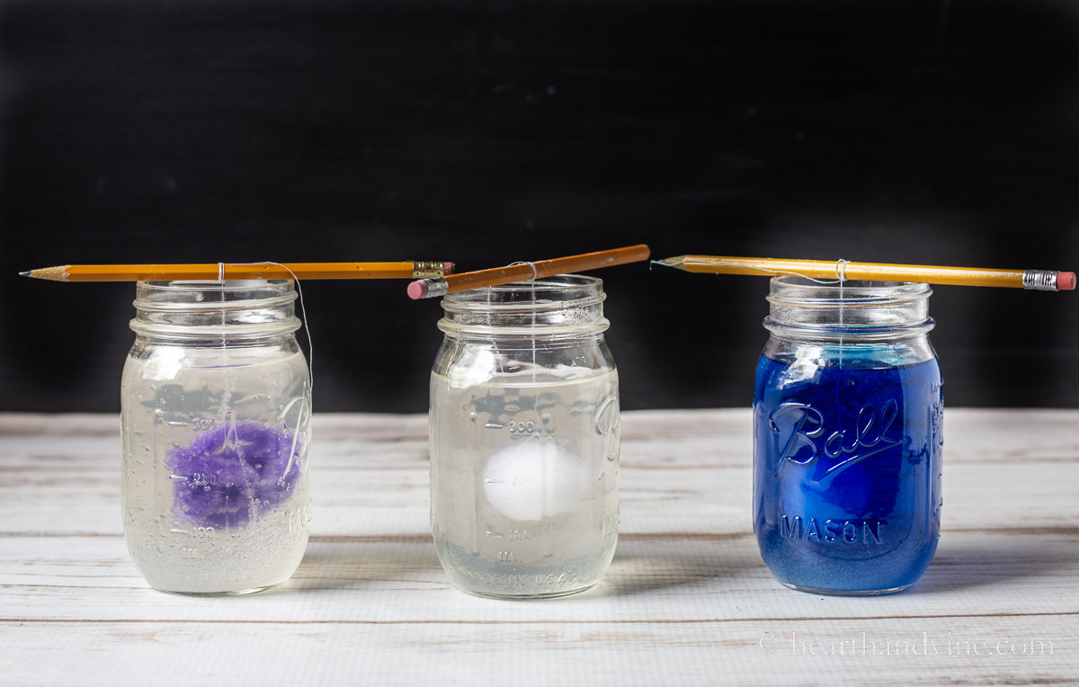 Three pint mason jars with pipe cleaners or pom poms suspended in borax and water solution by thread and pencil. One jar also has blue food coloring.