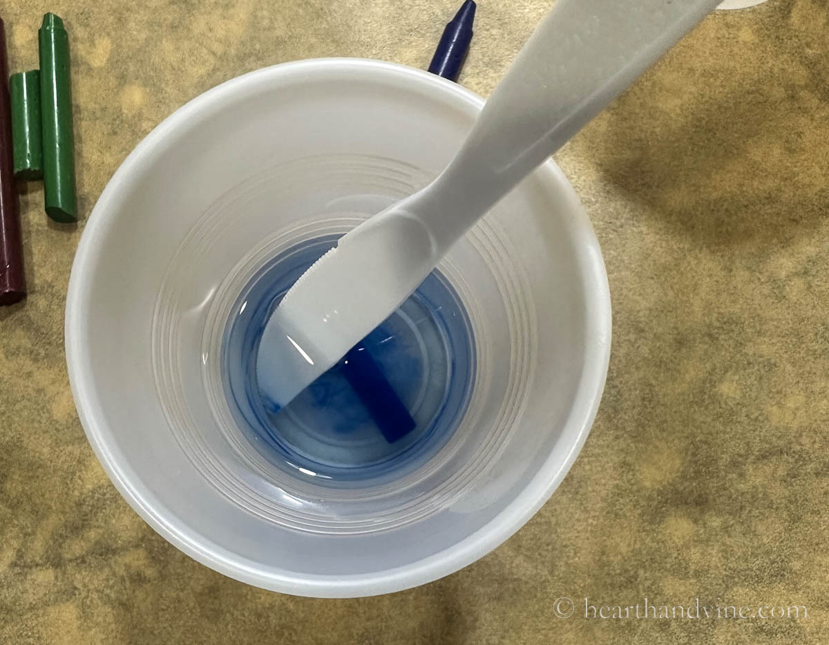 Piece of blue crayon melting in hot wax in a plastic cup with a plastic knife for stirring.
