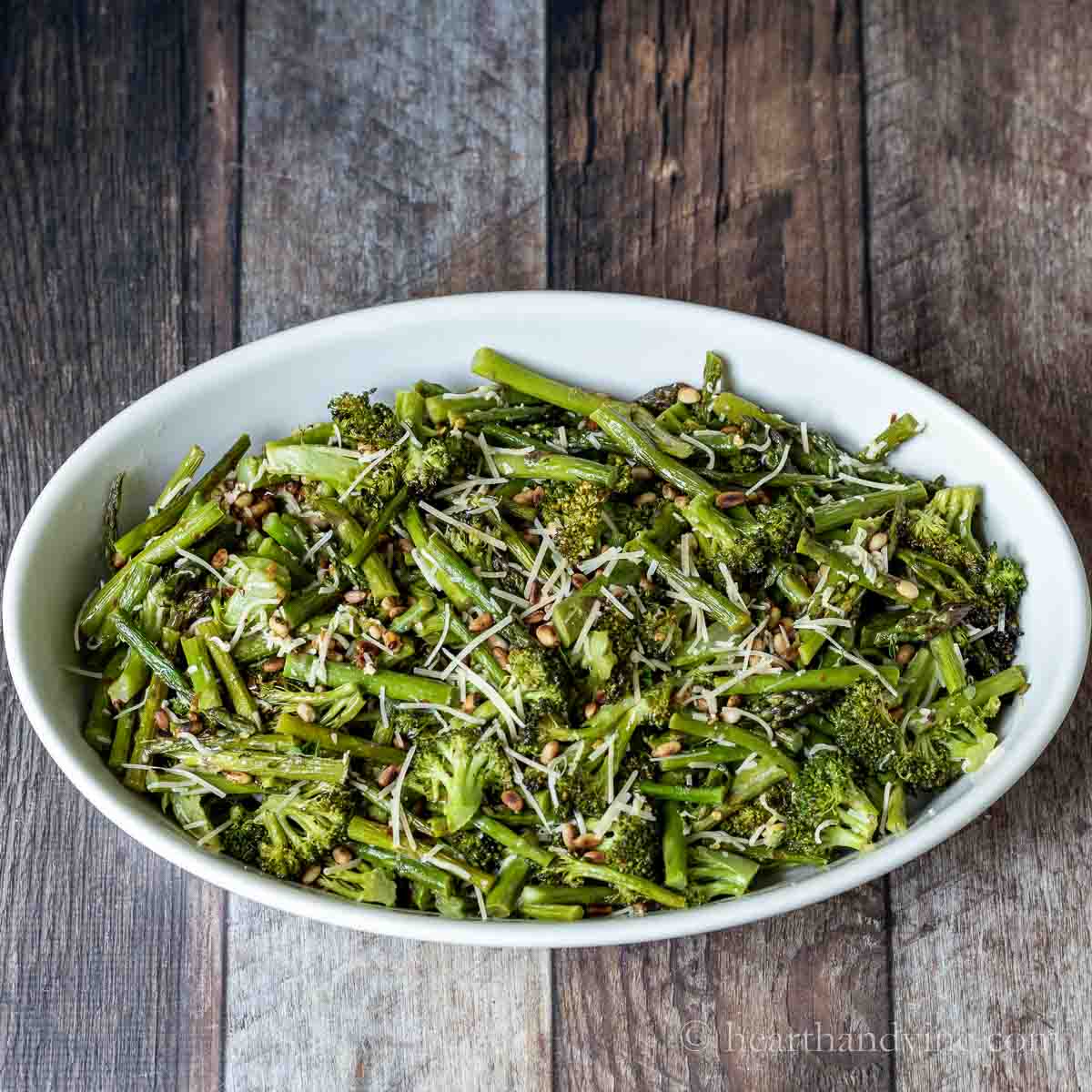 Oval baking dish with roasted broccoli and asparagus topped with cheese and pine nuts.