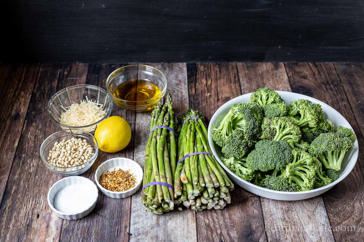 Ingredients including bunches of asparagus spears, broccoli florets, lemon, salt, pepper flakes, pine nuts and parmesan cheese.