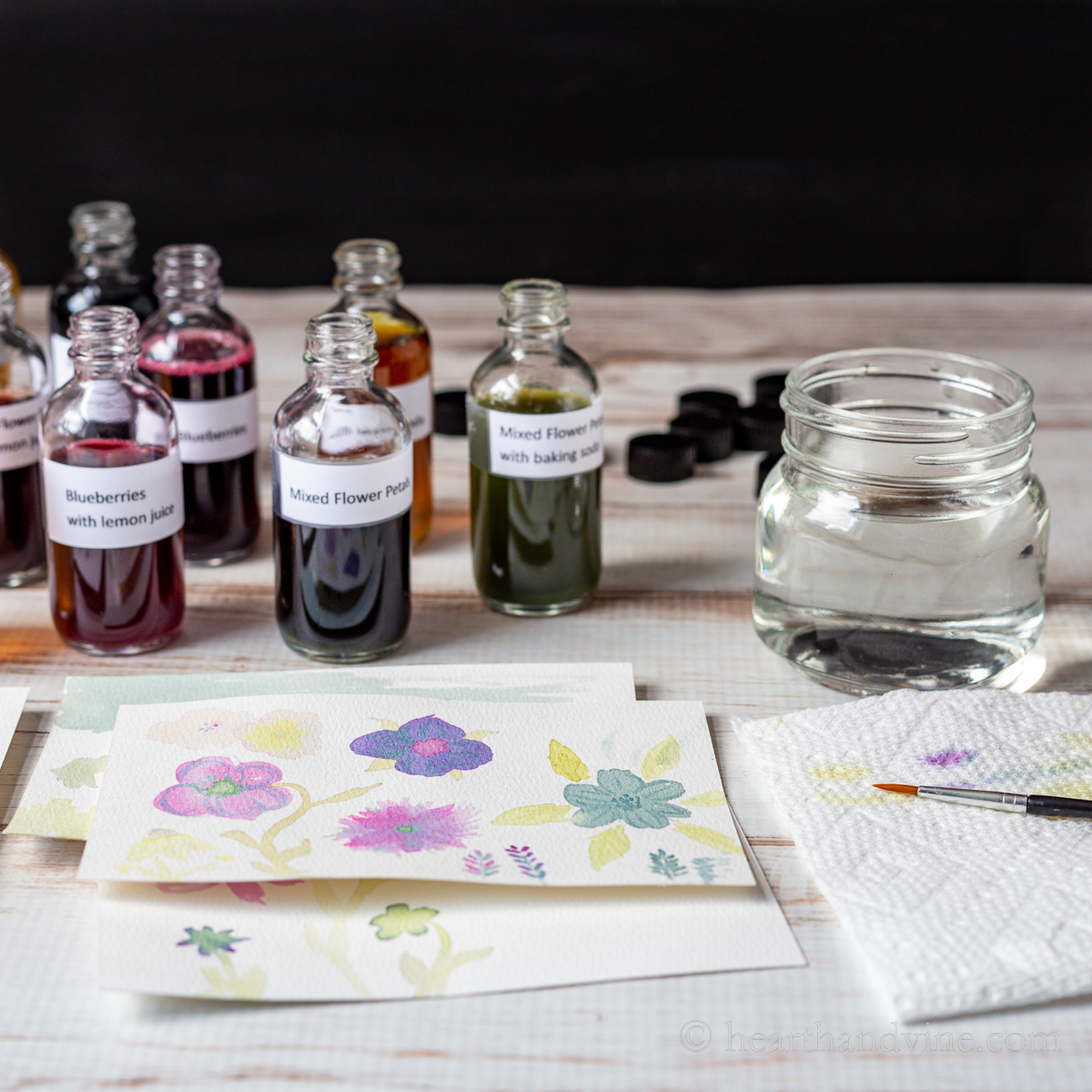 Labeled bottles of homemade watercolors and a jar of water, a couple of flower painting on small cards and a small paintbrush.