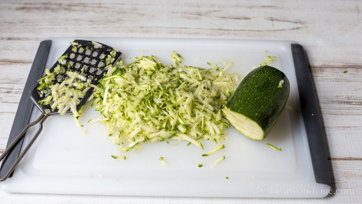 Grated zucchini, a hand grater and the bottom portion of a zucchini on a chopping board.