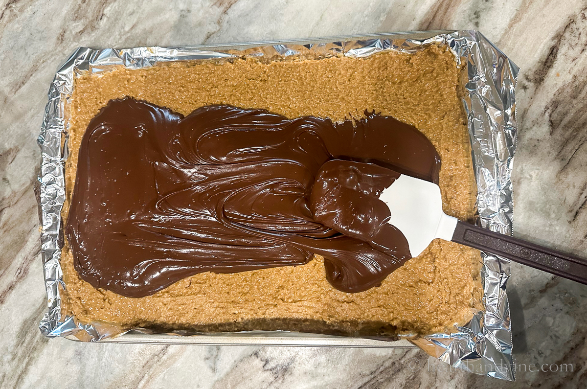 A rubber spatula spreading chocolate on top of the peanut butter mixture in the pan.