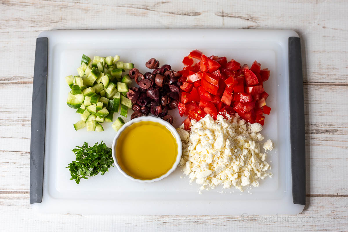 Chopping boards with chopped cucumbers, olives, tomatoes, parsley, crumbled feta cheese and a small bowl of olive oil.