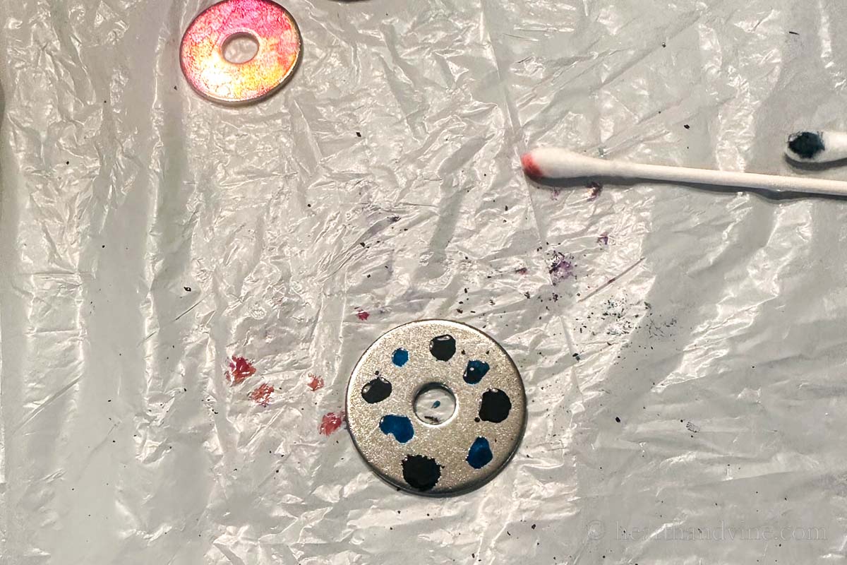 A metal washer with a few drops of alcohol ink next to a couple of cotton swabs and a painted washer.