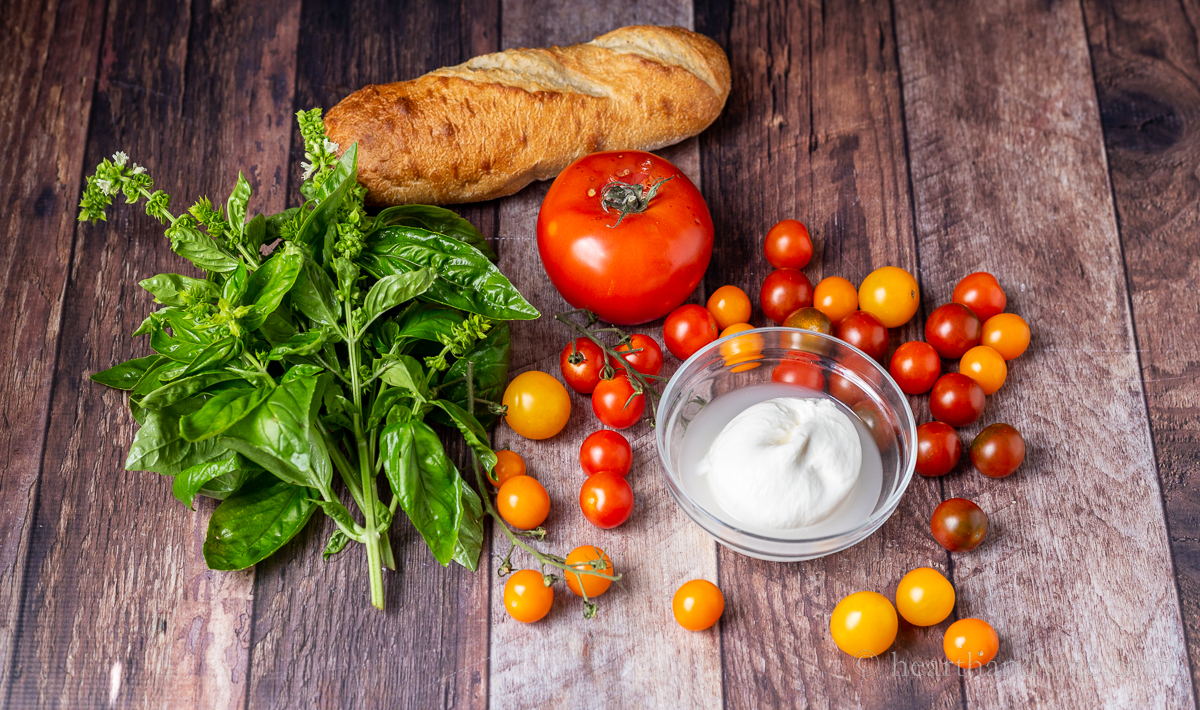Ingredients for burrata caprese including a burrata ball, fresh basil, a large tomato and heirloom cherry tomatoes, baguette bread.