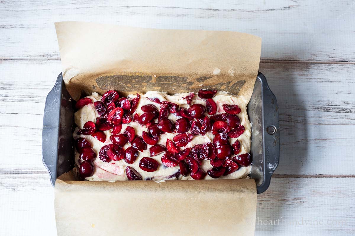 A loaf pan lined with parchment paper with batter and a layer of chopped cherries.