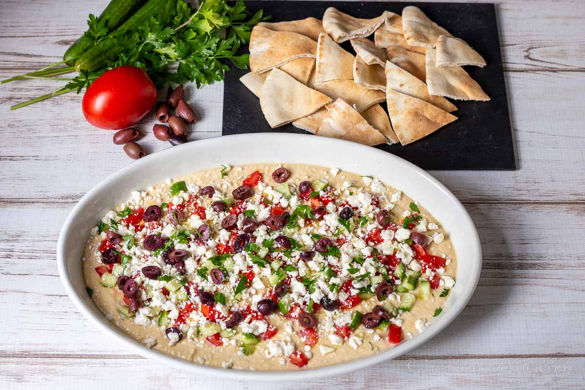 Greek layered hummus dip in an oval serving dish next to a tray of pita bread triangles, a Roma tomato, Kalamata olives, and parsely.