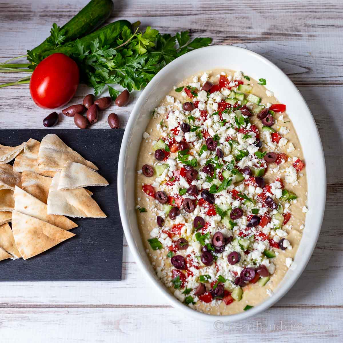 Oval serving dish with Greek layered hummus next to pita wedges, olives, tomato, cucumber, and parsley.