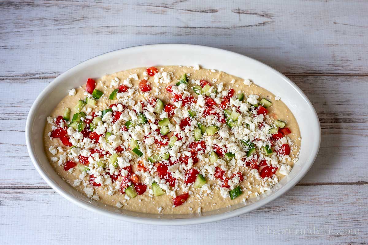 A large oval serving dish with a layer of hummus topped with chopped tomatoes, cucumbers, and crumbled feta cheese.