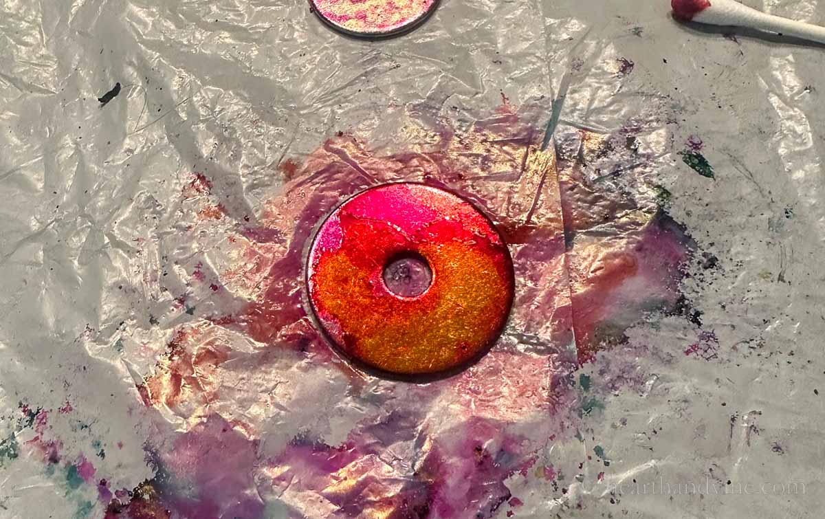 A metal washer with pink and yellow alcohol ink melding into each other.