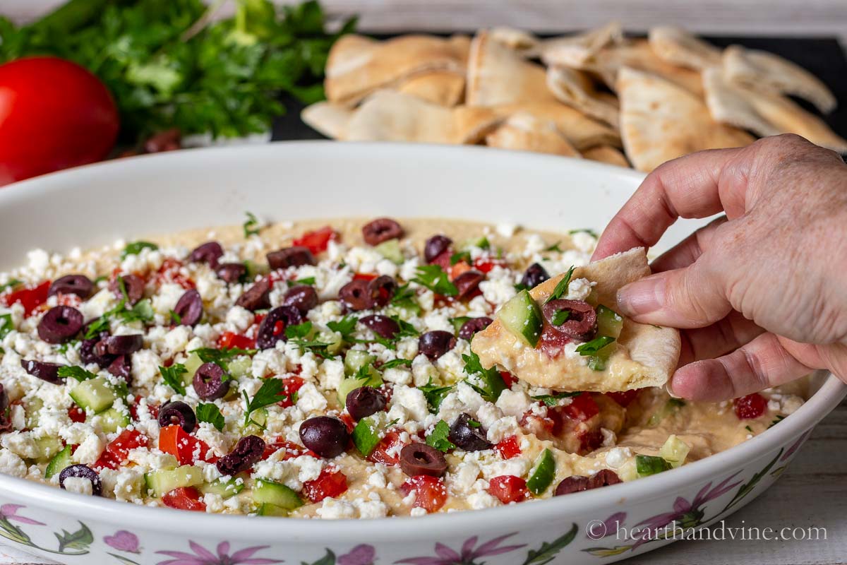 A hand with a pita bread wedge dipped into the Greek layered hummus dip.
