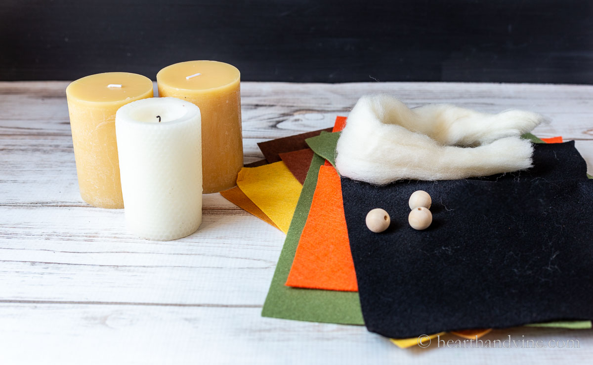 Supplies on a table including pillar candles, sheets of felt, wood beads and roving wool.