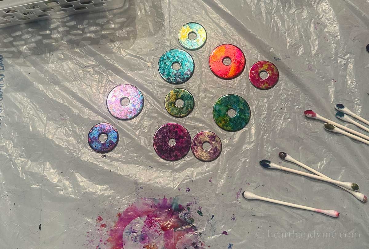 Several painted washers with alcohol inks and used cotton swabs on a plastic covered table.