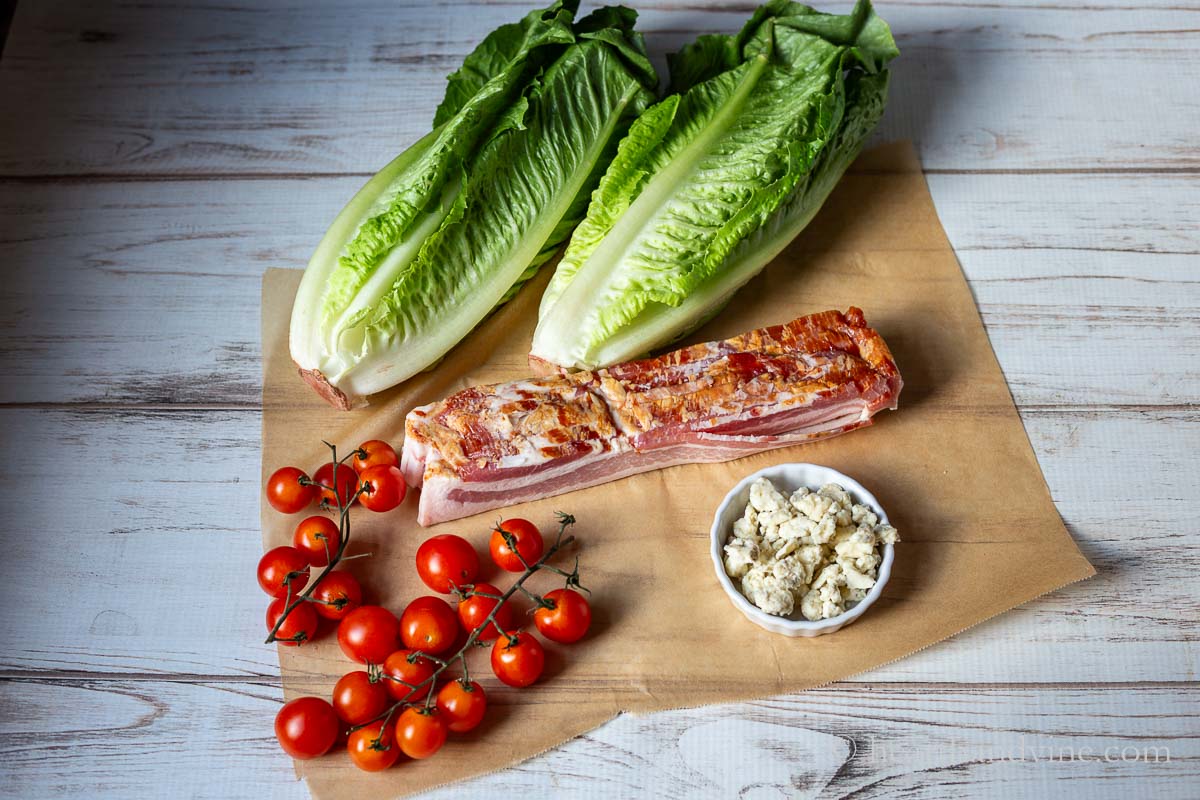 BLT salad ingredients including heads of romaine lettuce, cherry tomatoes, thick bacon slices and crumbled Gorgonzola cheese.