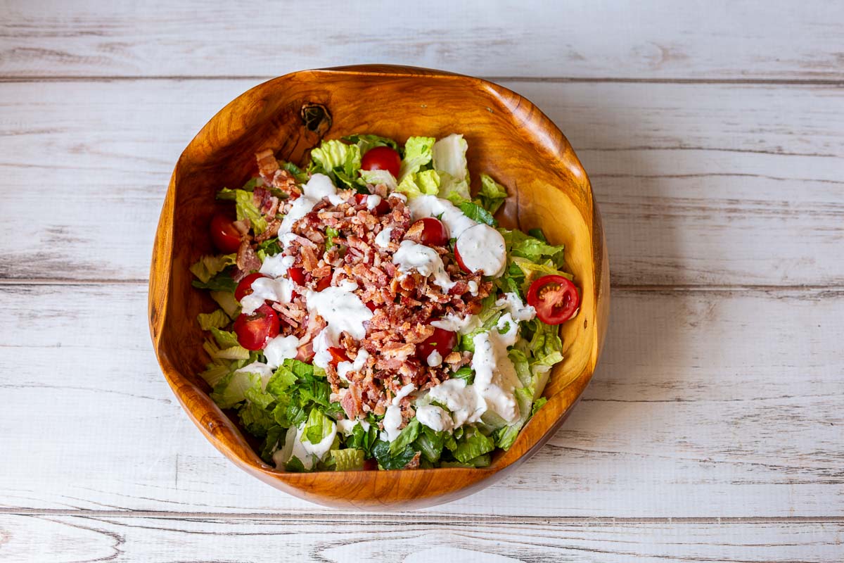 A large wooden salad bowl with romaine lettuce, cherry tomatoes, bacon and blue cheese dressing not tossed.
