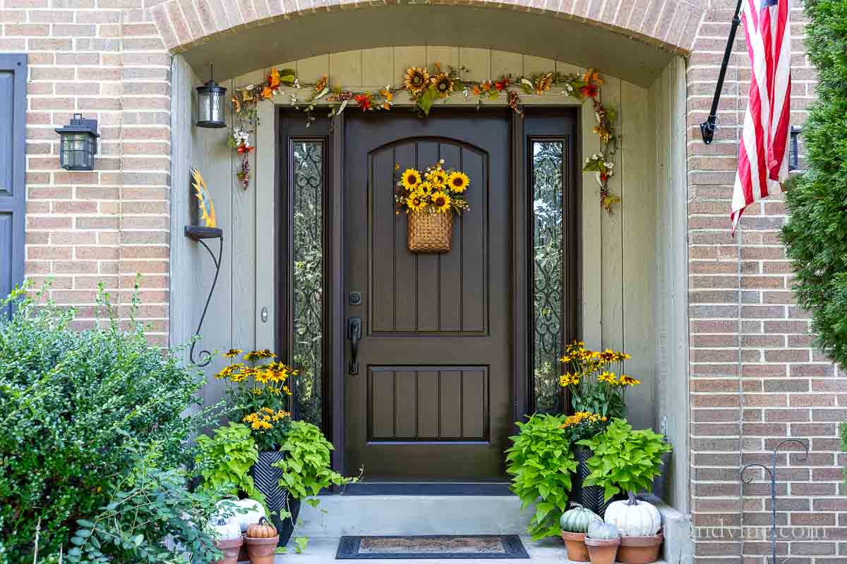 Front porch decorated for fall with a garland over the door, a sunflower basket on the door and fall flowers in pots surrounded by faux pumpkins in clay pots.