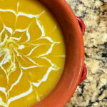 Partial view of a bowl of delicata squash soup with a spider web in cream design on top.