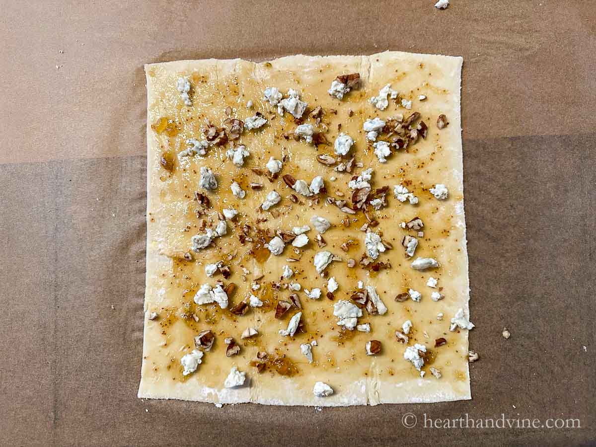 Puff pastry sheet with fig jam spread on top and blue cheese crumbles and chopped pecans scattered around.