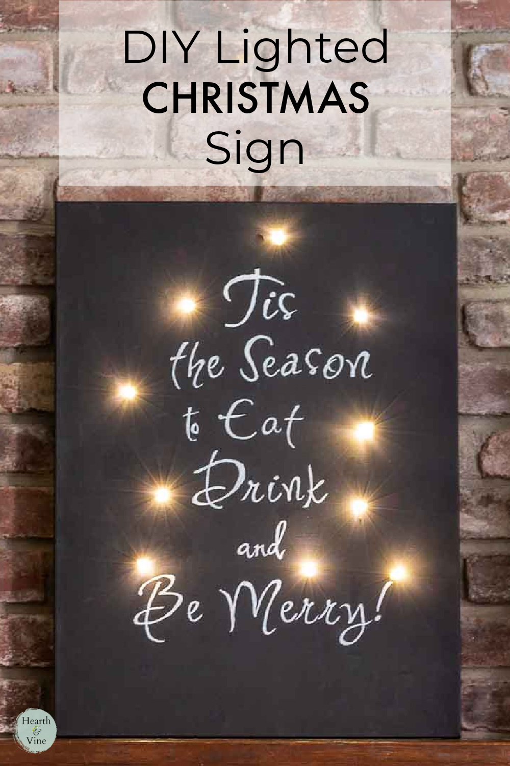 A black chalkboard look canvas with a white saying, "Tis the Season to Eat Drink and Be Merry" with LED lights around the edge.