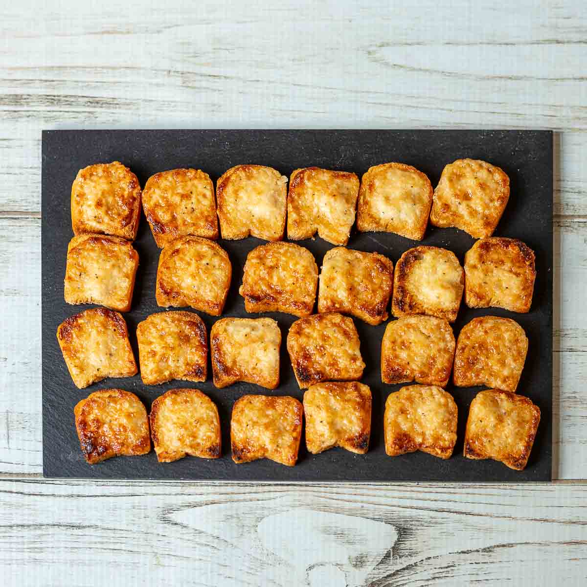 Parmesan bread bites on a black serving slate all lined up in rows.