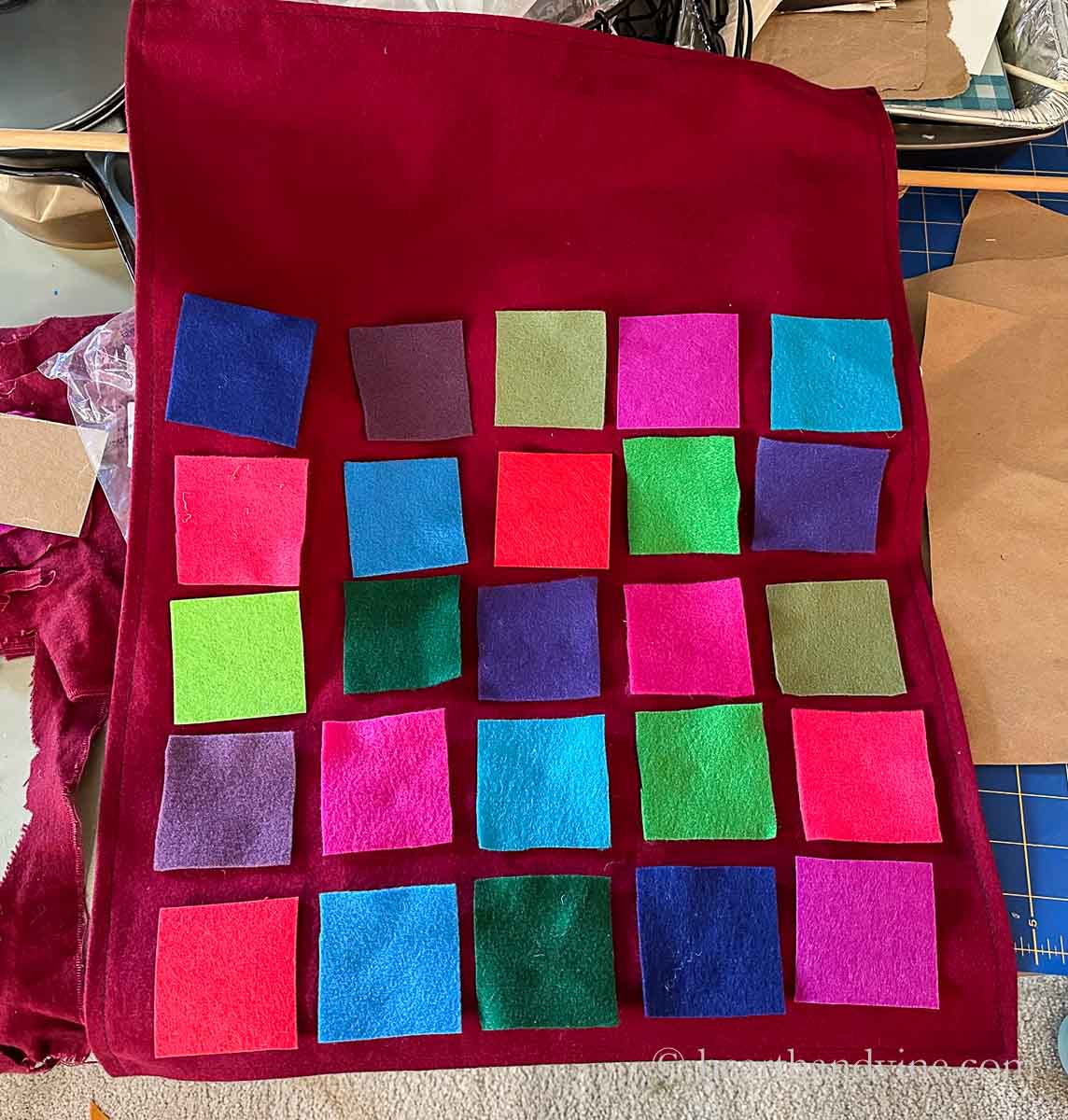Twenty five colored squares of felt arranged on a dark red backing.