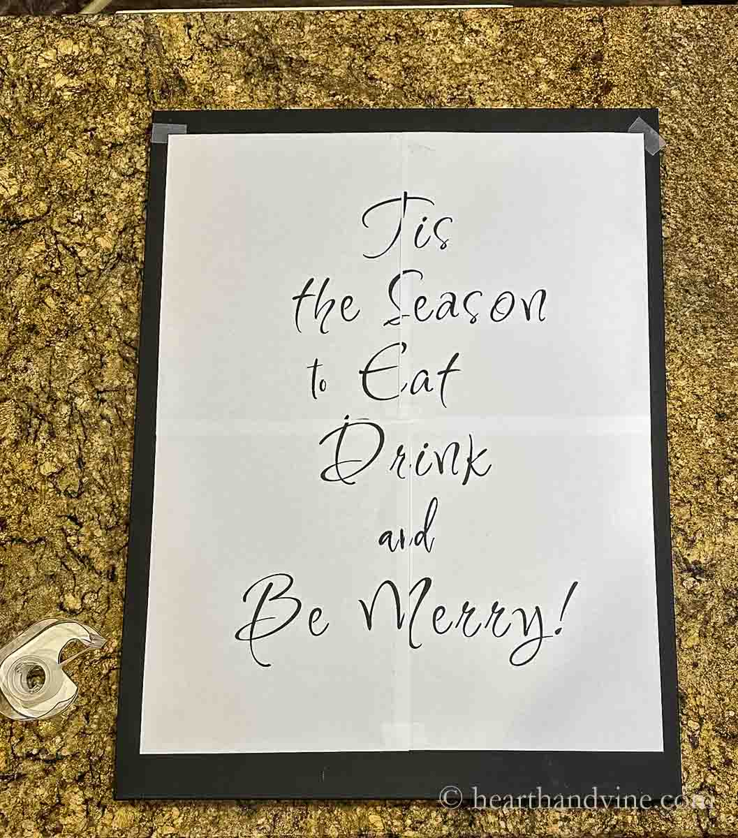 Four pages of computer paper printed out to create a large sign saying Tis the Season to Eat Drive and Be Merry, taped onto the black painted canvas.