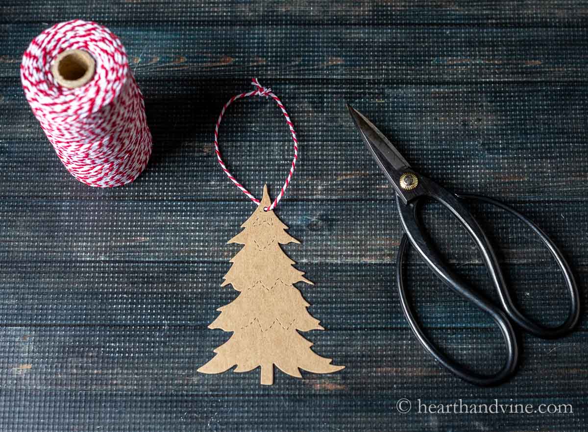 A spool of red and white twine next to a chipboard Christmas tree with a twine loop at the top and a pair of scissors.