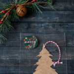 Chipboard tree in a bag with a Merry sticker next to a rusty bell and berry pine decoration.