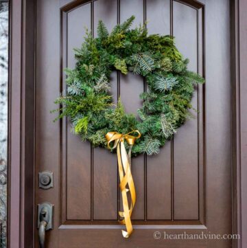 Christmas wreath with different kinds of greenery on a front door with a gold ribbon.