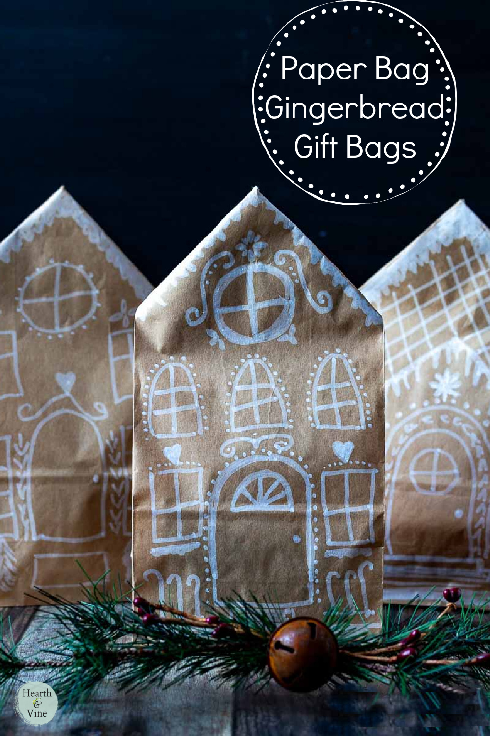 Three paper bag gingerbread house gift bags with a small piece of faux pine, berries and a rusty bell in front.