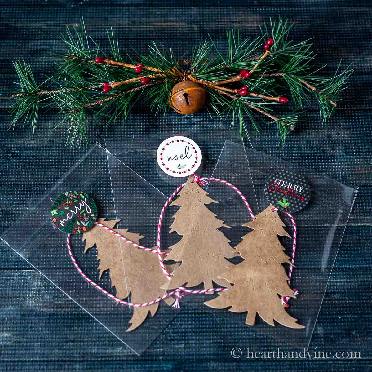Three bagged chipboard trees with Christmas labels next to a bell with pine decoration.