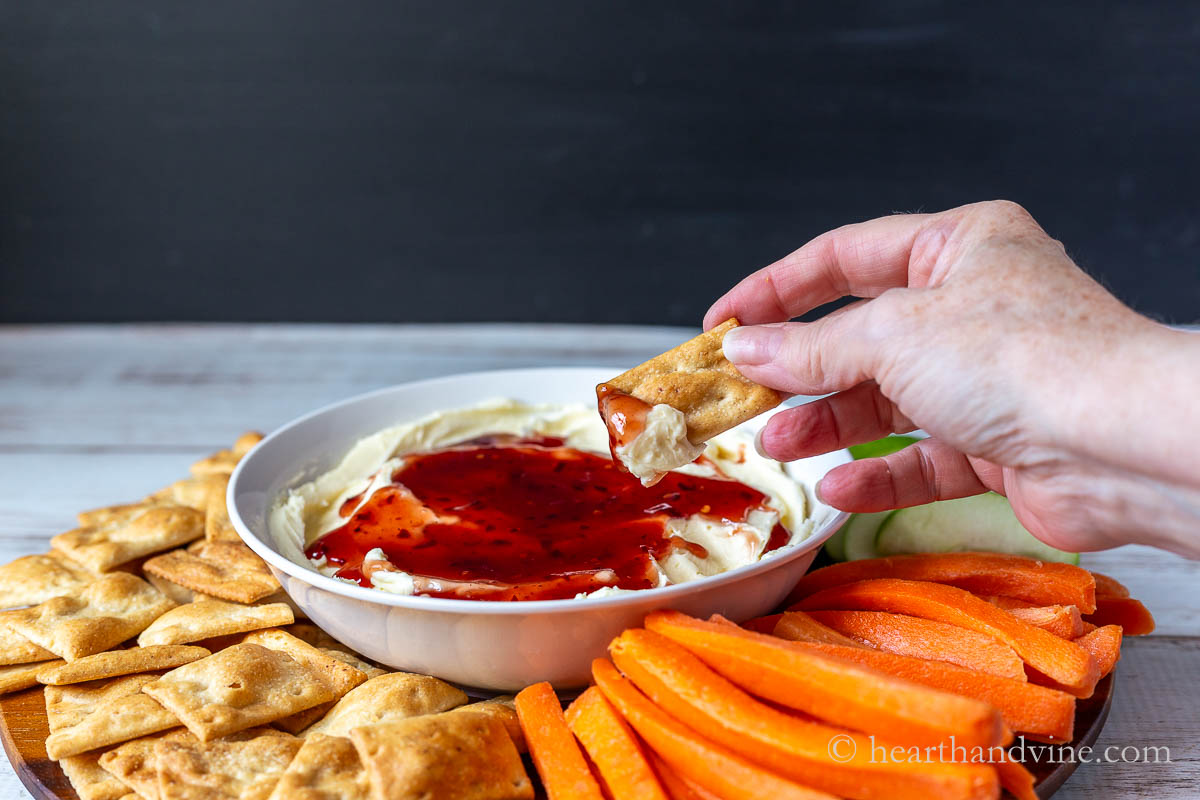A hand with a cracker dipping into the whipped brie dip with some jam.