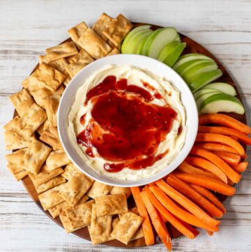 A bowl of whipped brie dip with raspberry jam surrounded by crackers, carrots and apple slices.