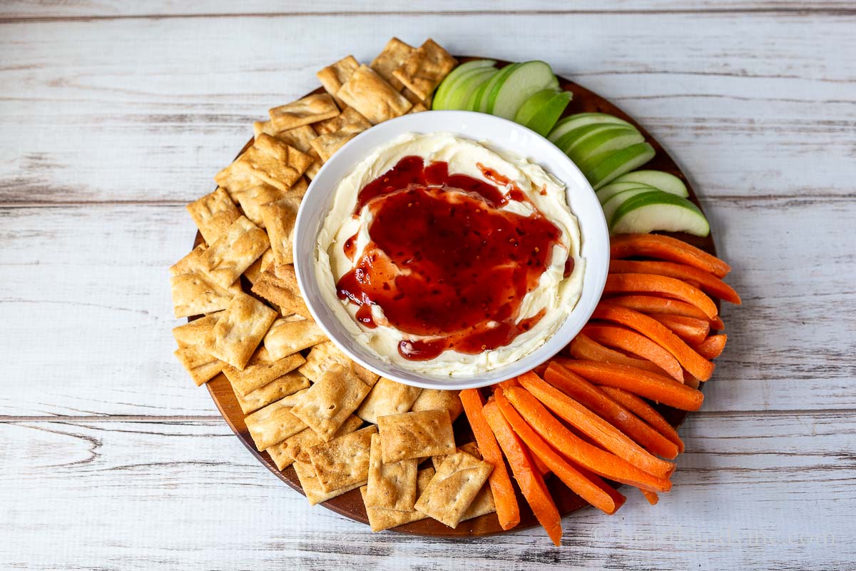 A round platter with green apple slices, crackers, and carrot sticks surrounding a bowl of whipped brie with raspberry jam.