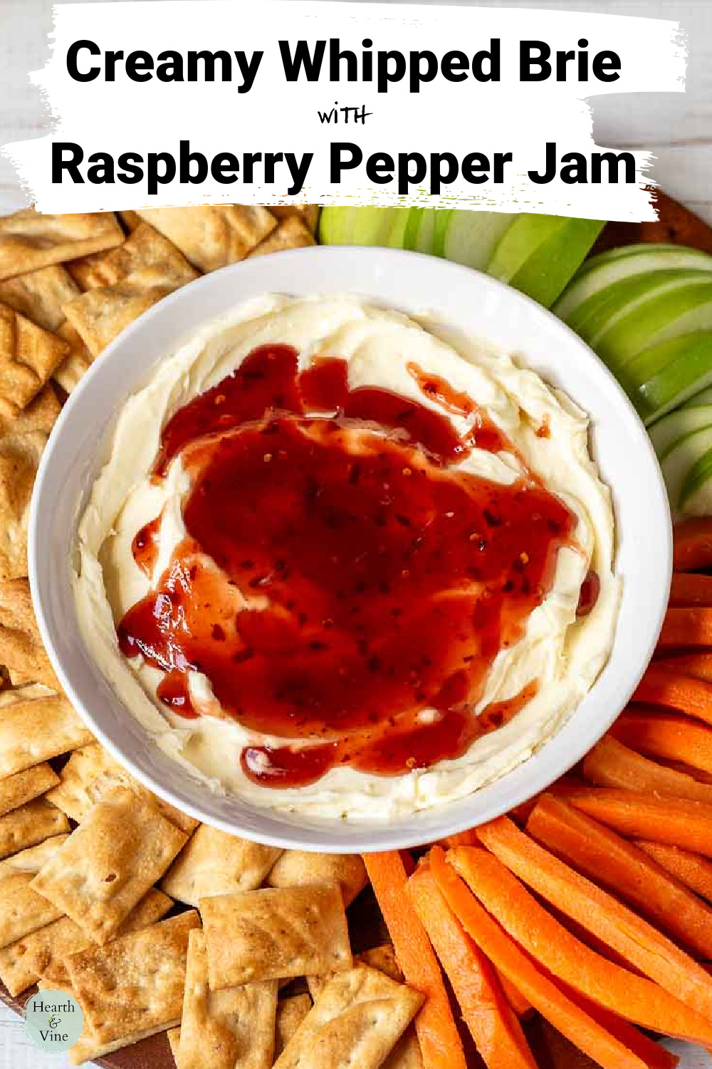 White bowl filled with whipped brie and topped with pepper raspberry jam on a platter with crackers, carrot sticks and green apple slices.