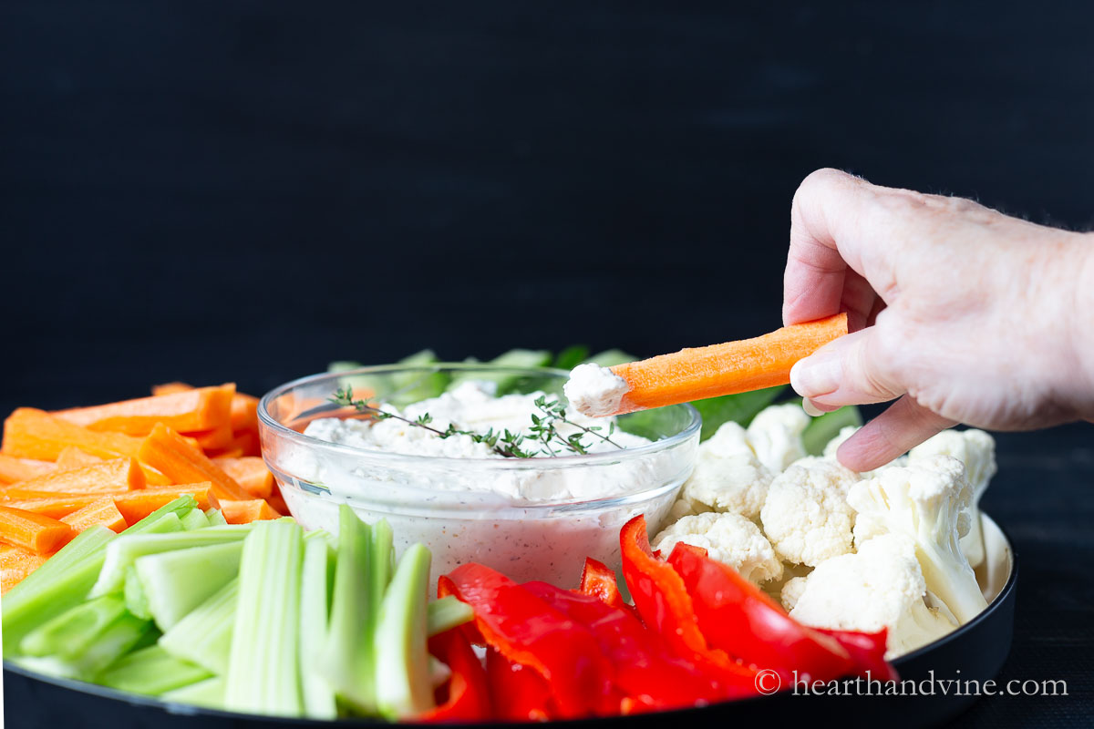 A hand holding a carrot stick that has been dipped into a cream cheese dip over a platter with the dip and other vegetables.