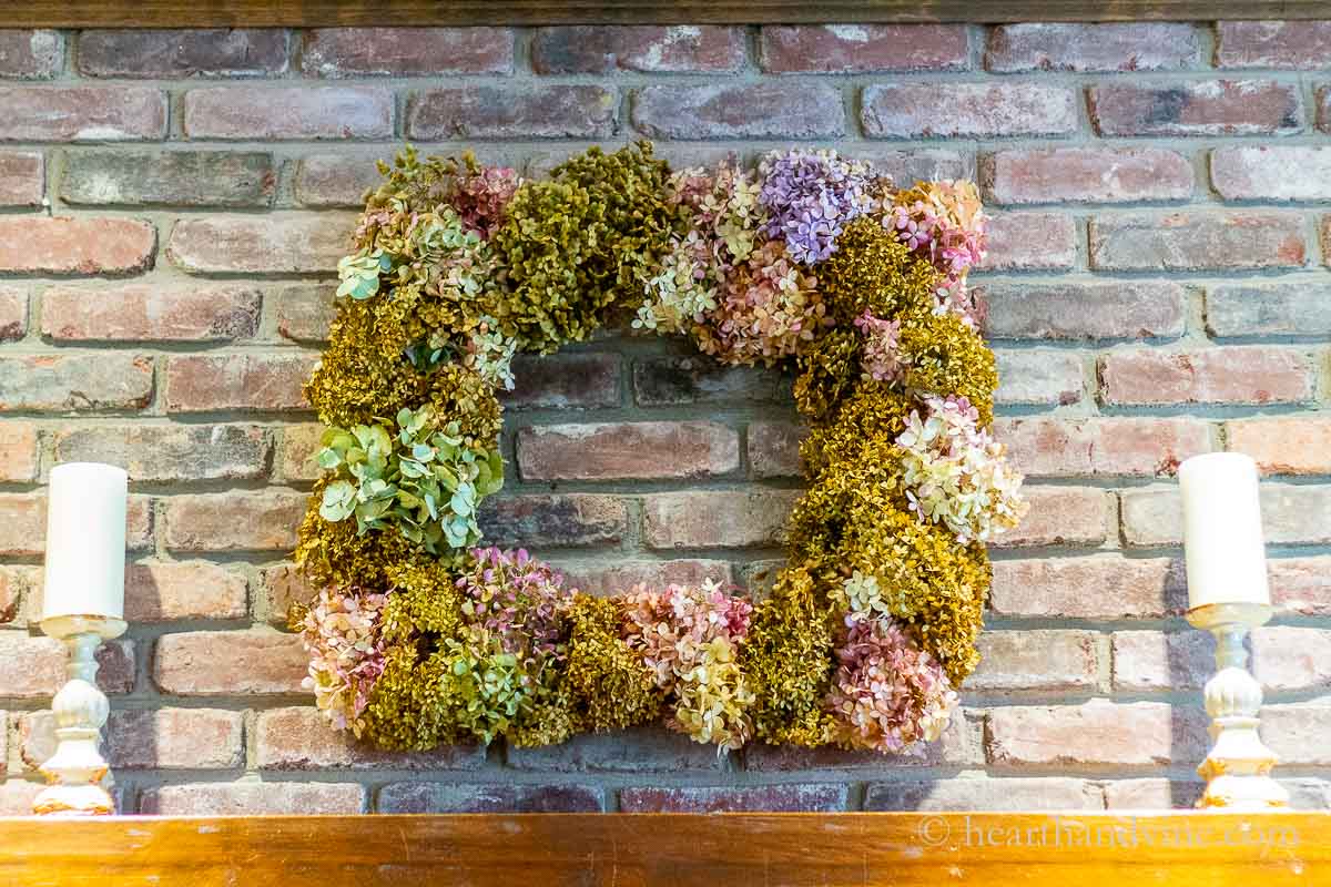 Square dried hydrangea wreath hanging above a mantel on a brick wall.