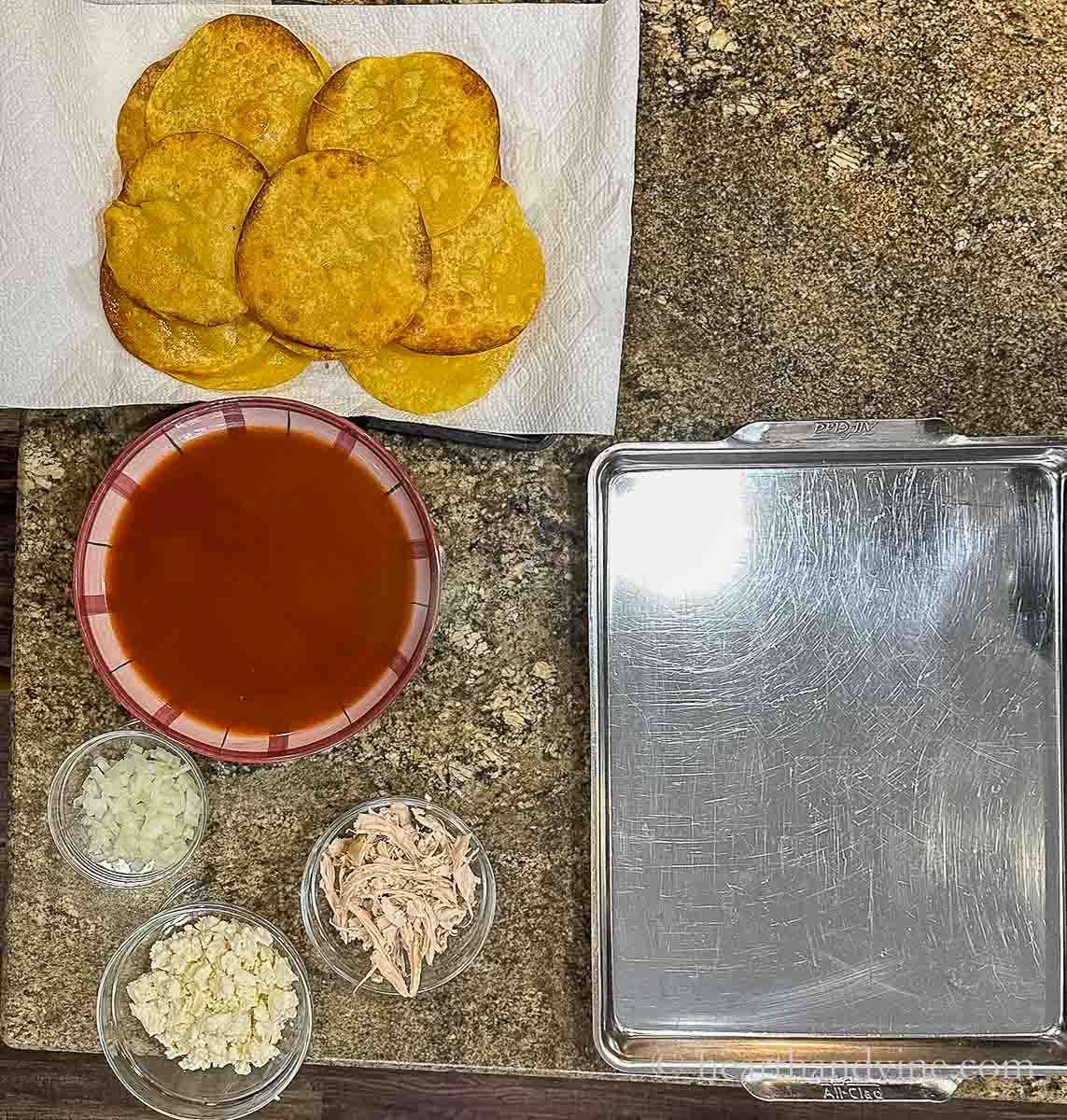 Fried corn tortillas, a bowl of red enchilada sauce, bowls of chopped onions, queso fresco and shredded chicken.