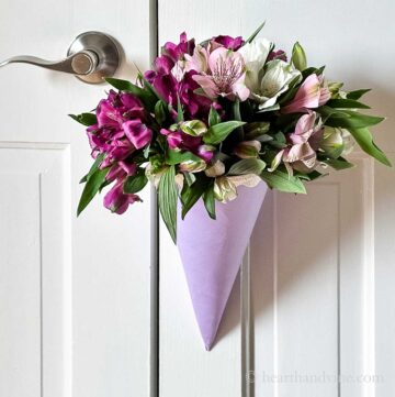 A lavender paper flower cone with pink ribbon edge holding a bouquet of flowers hanging on a door.