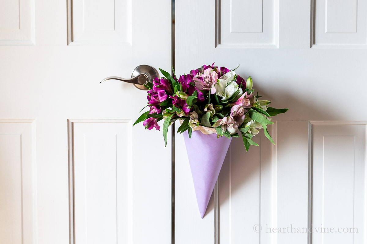 A lavender paper cone with pink ribbon edging holding a large bouquet of flowers hanging on a white door.